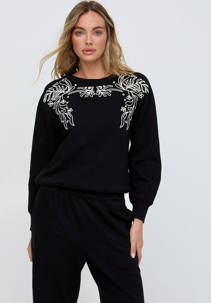 Woman wearing a black outfit with white floral embroidery on a plain background