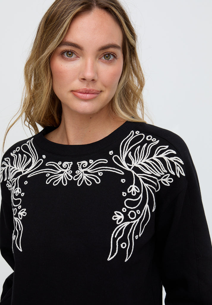 Woman wearing a black sweater with white floral embroidery on a plain background