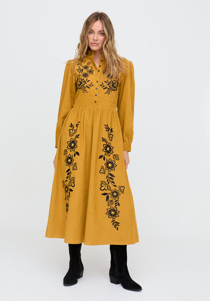 Woman wearing a mustard yellow dress with black floral embroidery on a white background