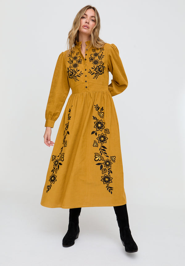 Woman wearing a mustard yellow dress with black floral embroidery on a white background