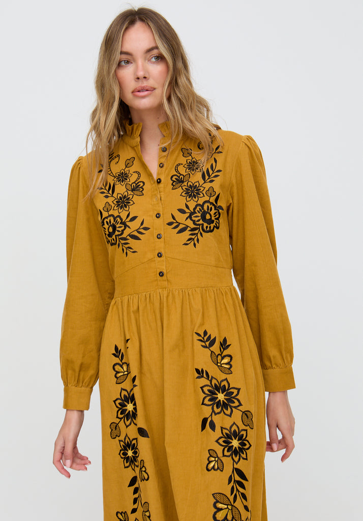 Woman wearing a mustard yellow dress with black floral embroidery on a white background