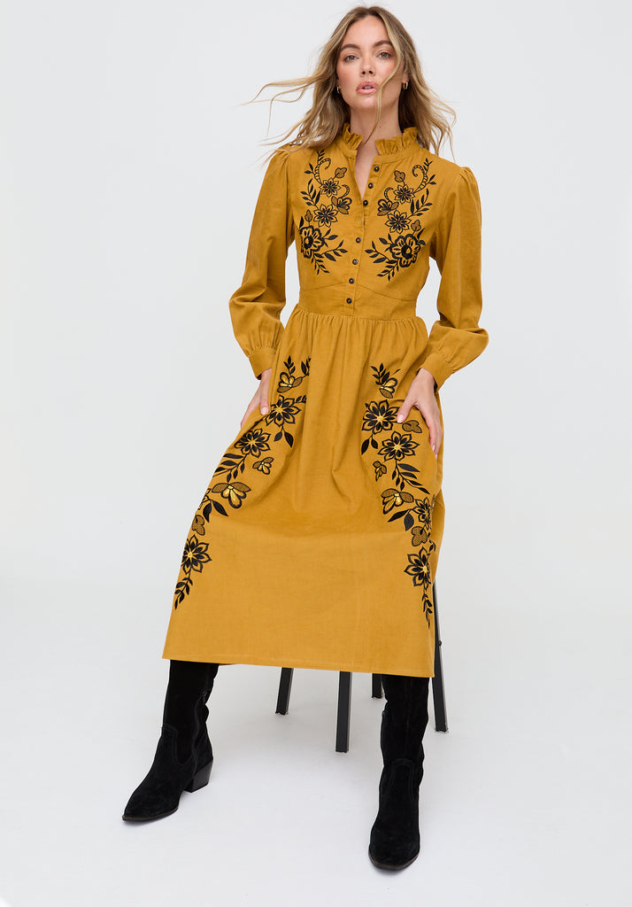 Woman wearing a mustard yellow dress with black floral embroidery on a white background