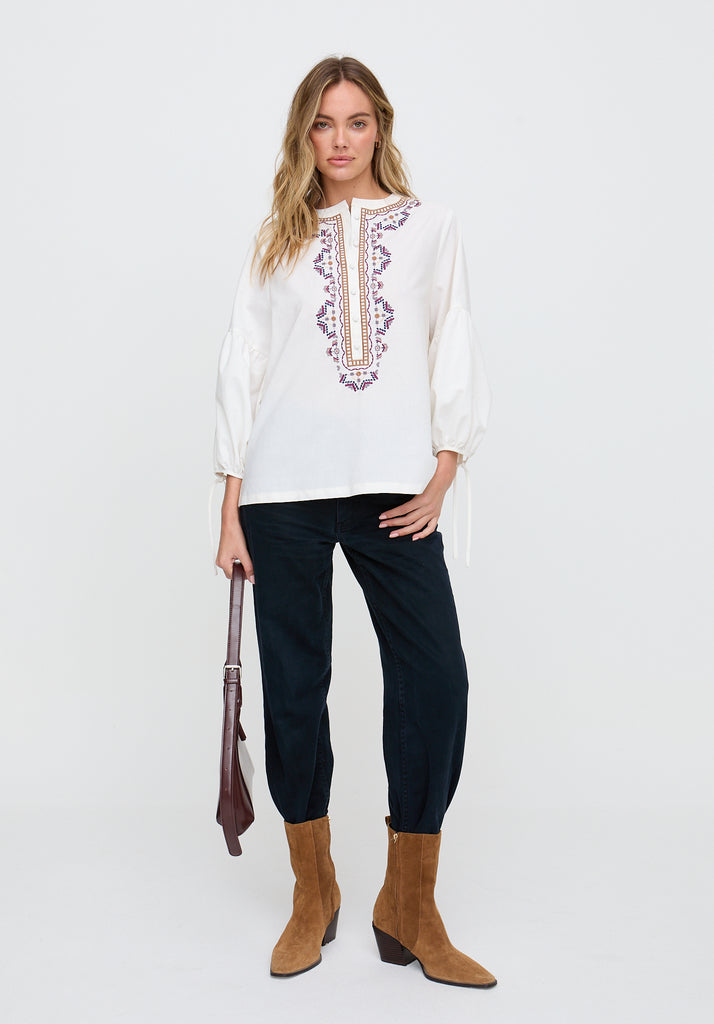 Woman wearing a white embroidered blouse with black pants and brown boots on a white background