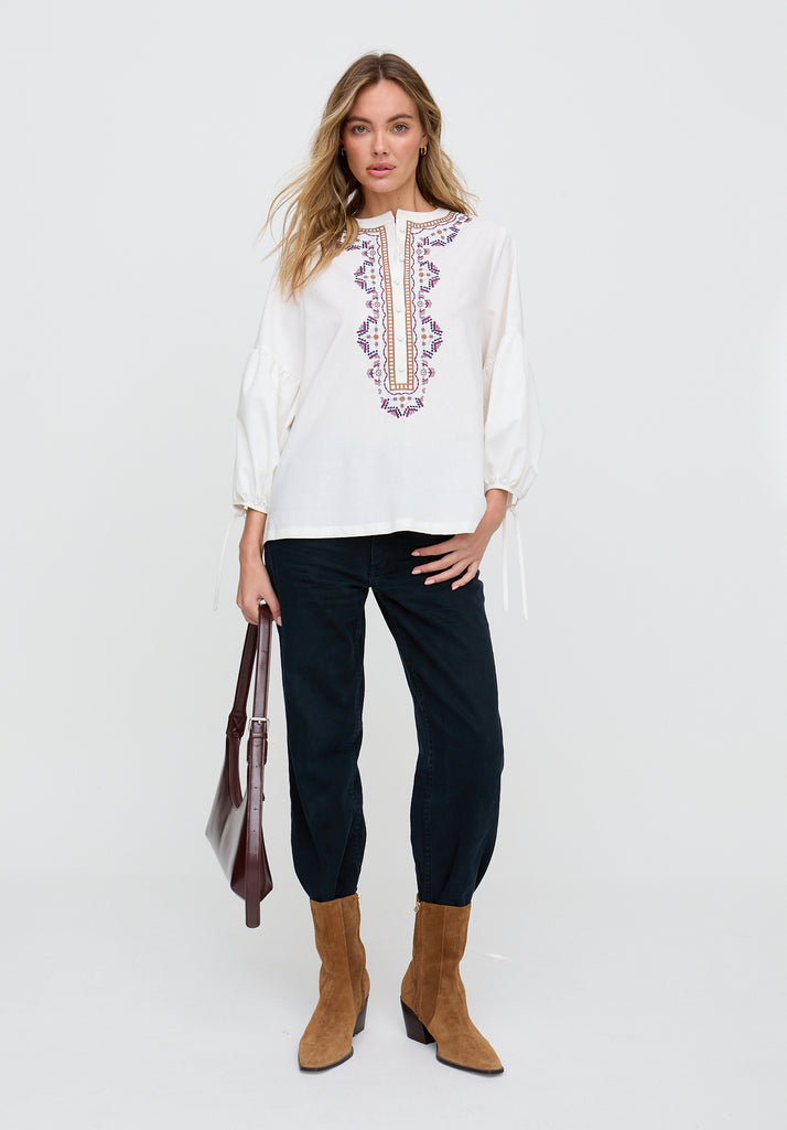 Woman wearing a white embroidered blouse with black pants and brown boots on a white background