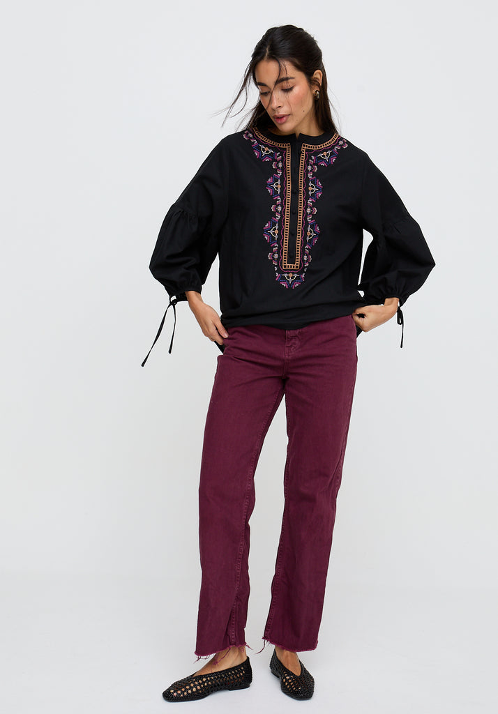 Woman wearing a black blouse with purple embroidery and burgundy pants on a white background