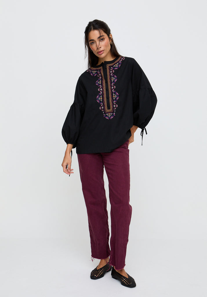 Woman wearing a black embroidered top and burgundy pants on a white background