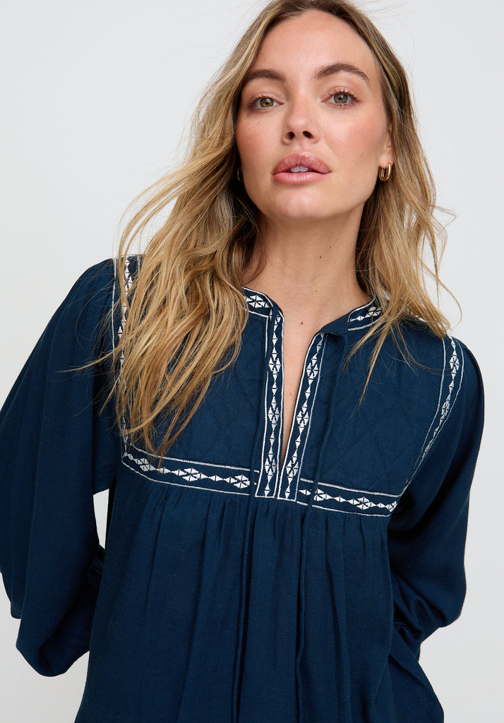 Woman wearing a navy blue dress with white embroidery on a plain background