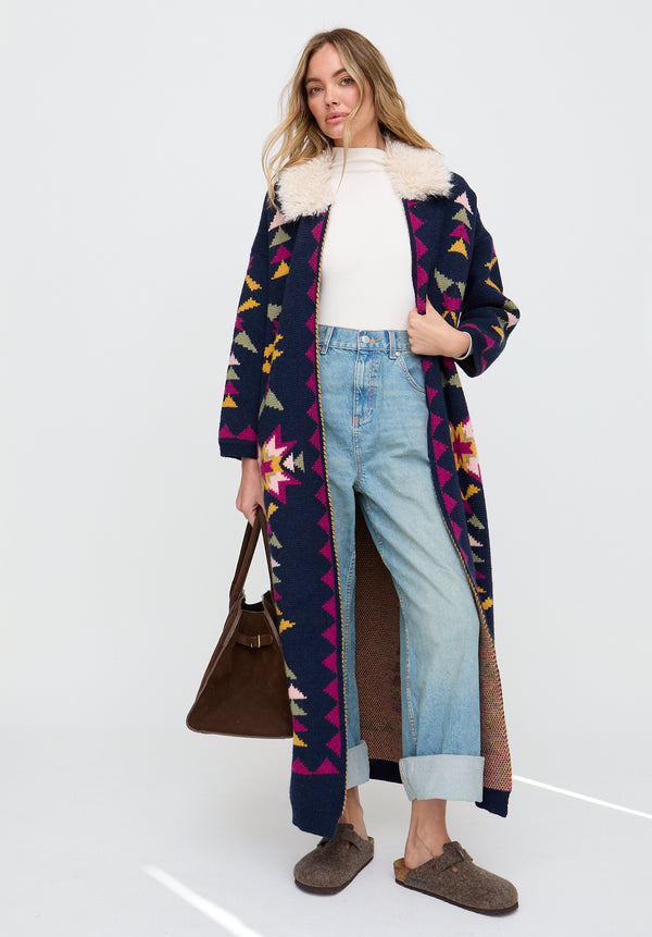 Woman wearing a long, patterned coat over a white sweater and jeans, holding a brown bag on a white background.