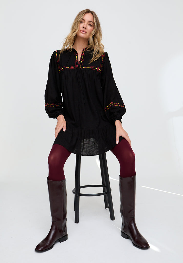 Woman wearing a black long-sleeve top with red embroidery, black leggings, and brown knee-high boots on a white background.