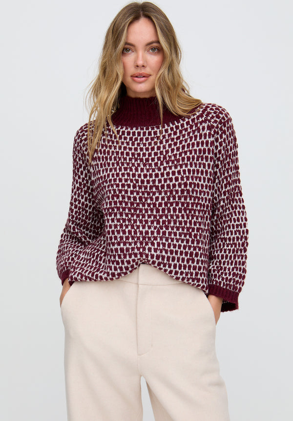 Woman wearing a patterned sweater and beige pants against a plain background