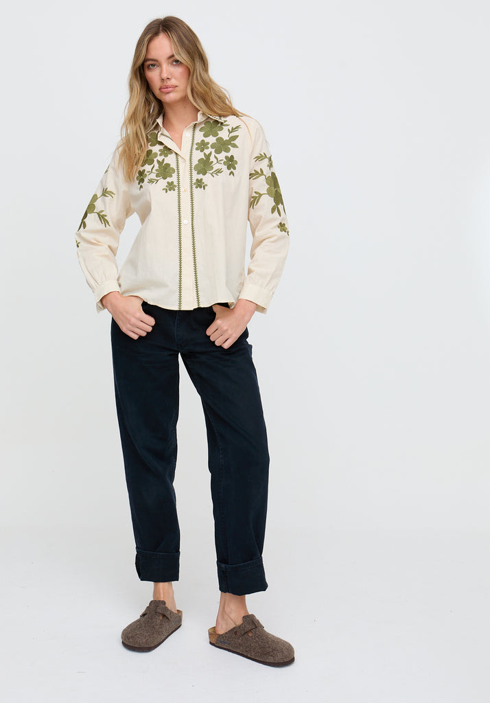 Woman wearing a cream floral blouse and dark pants on a white background