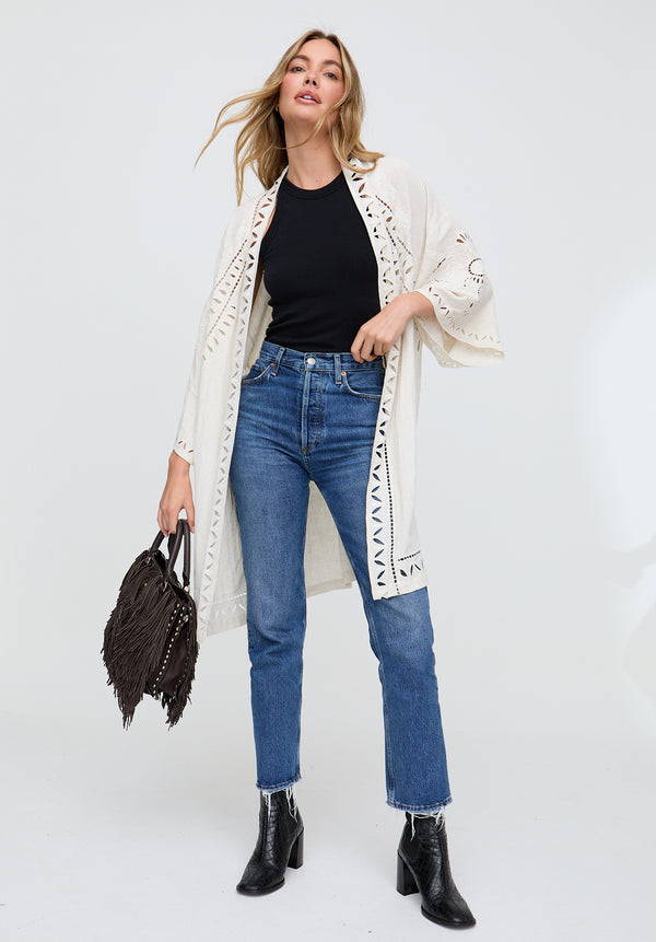 Woman wearing a long white cardigan, black top, blue jeans, and black boots on a white background