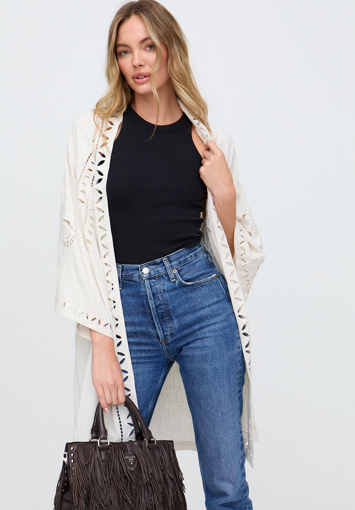 Woman wearing a white lace cardigan, black top, blue jeans, and holding a brown handbag on a light gray background