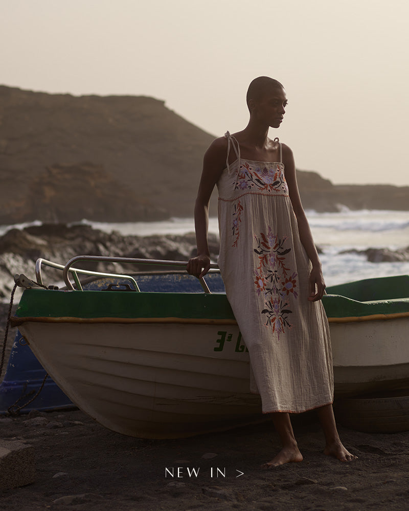 Model on beach, leaning on boat. Model is wearing a floral embroidered midi dress. Copy on image 'New In'