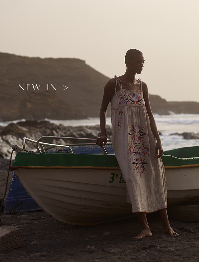 Model on beach, leaning on boat. Model is wearing a floral embroidered midi dress. Copy on image 'New In'