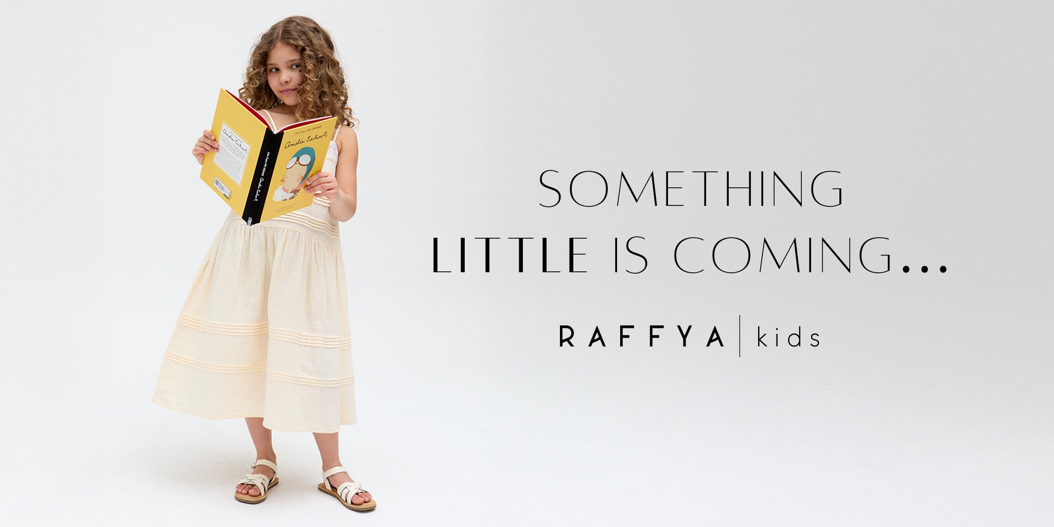 Raffya Kids | Girls stands reading emelia earhart book whilst wearing raffya kids linen cream midi dress with embroidered detailing. 