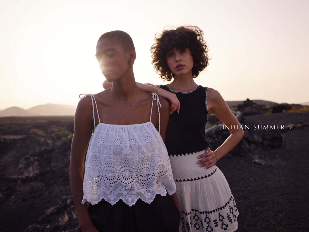 Model on left wearing white embroidered cami with tie detail on shoulders. Model on right wearing black sleeveless jersey top with stitching detail, and a cream mini skirt with black geometric embroidery. Copy on image 'Indian Summer'