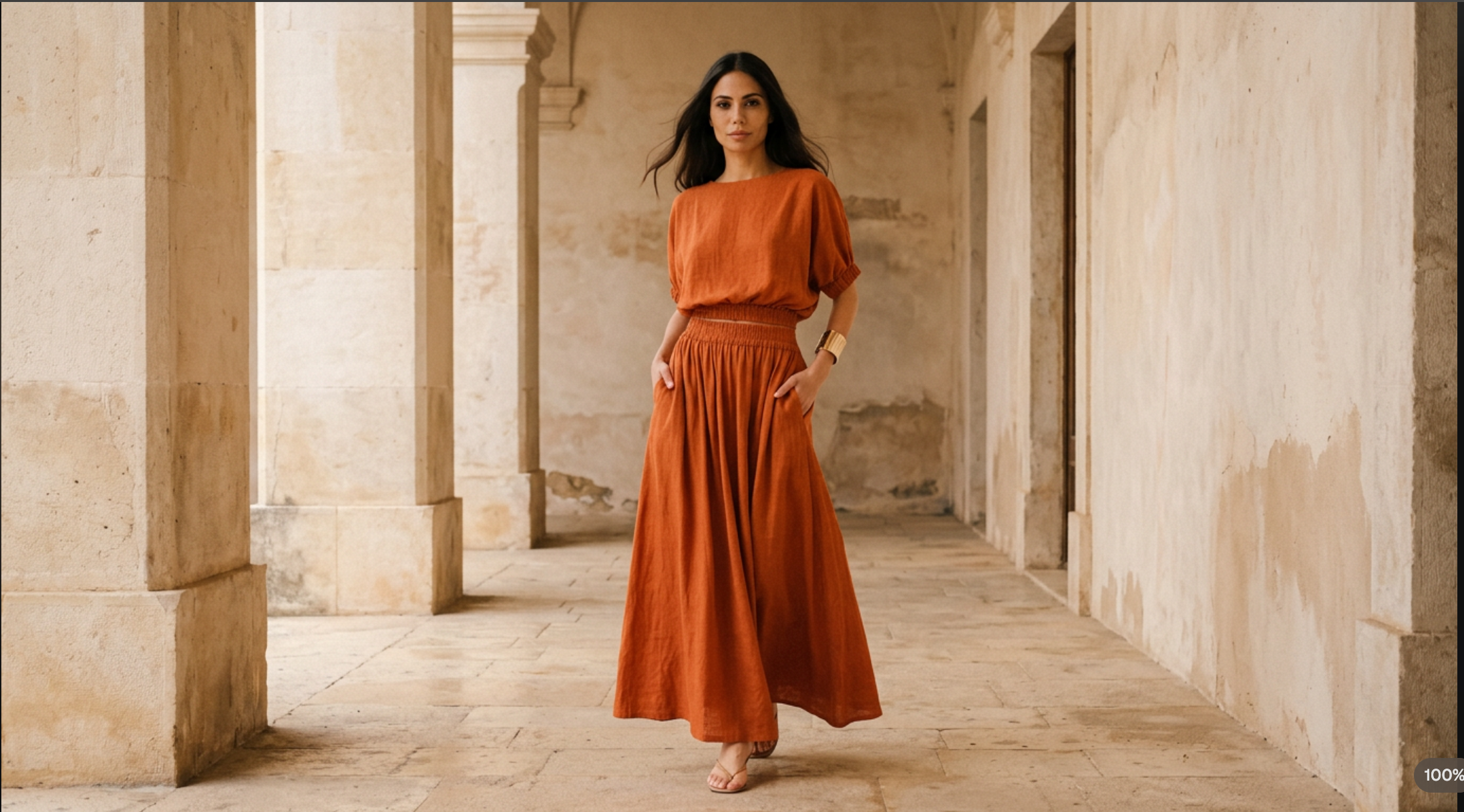Woman in an orange dress standing in a sunlit hallway with stone walls.